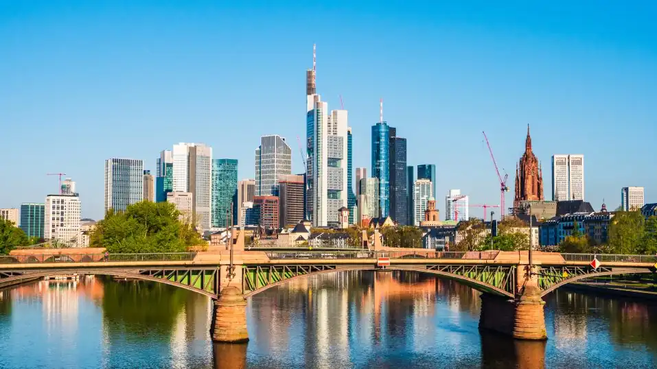 die Skyline der City Frankfurt unter blauem Himmel, im Vordergrund der Main mit Brücke