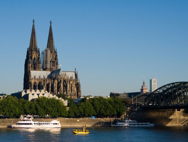 Foto Der Kölner Dom unter blauem Himmel, vorn der Rhein mit Schiffen, rechts die Hohenzollernbrücke