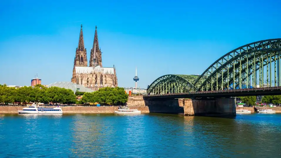 Der Kölner Dom unter blauem Himmel, vorn der Rhein mit Schiffen, rechts die Hohenzollernbrücke